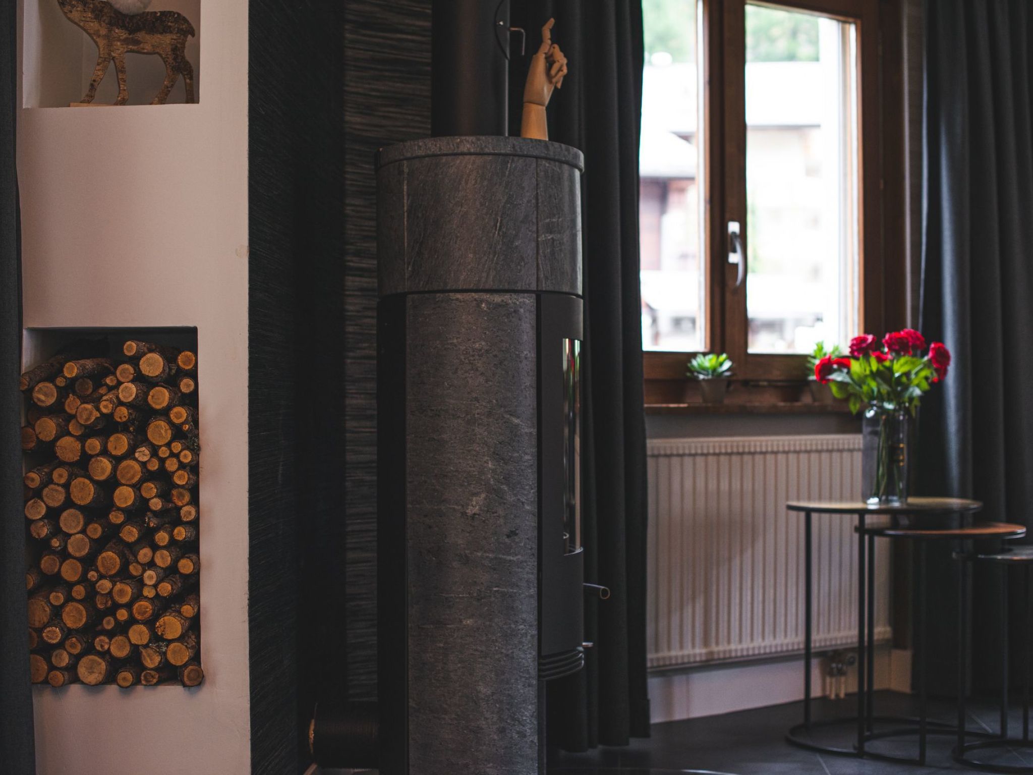 Een kamer met een open haard, een houtopslag aan de muur, een raam met gordijnen en een kleine tafel met een bloemenvaas.