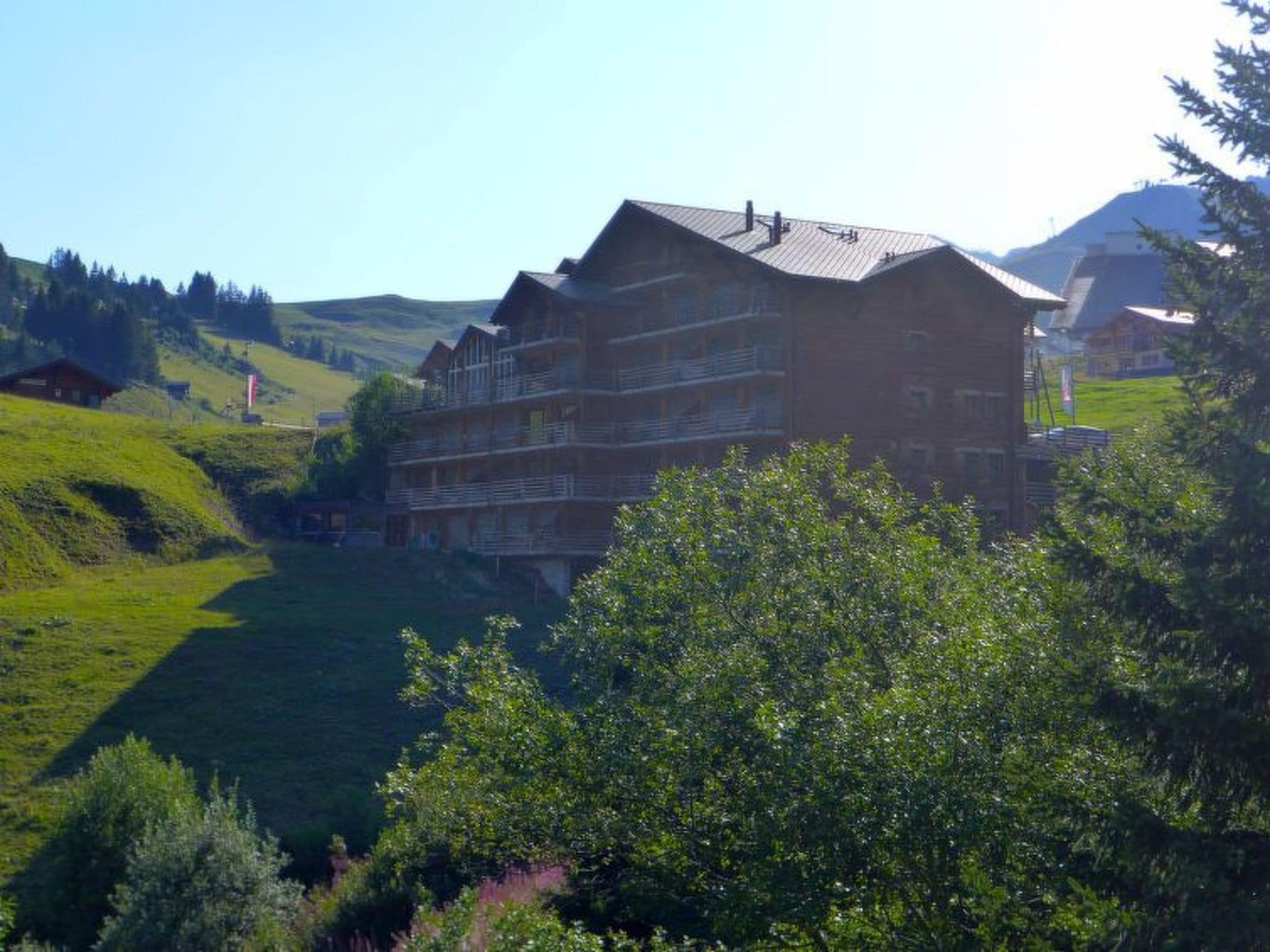 A multi-story building with a sloped roof, surrounded by lush greenery and hills.
