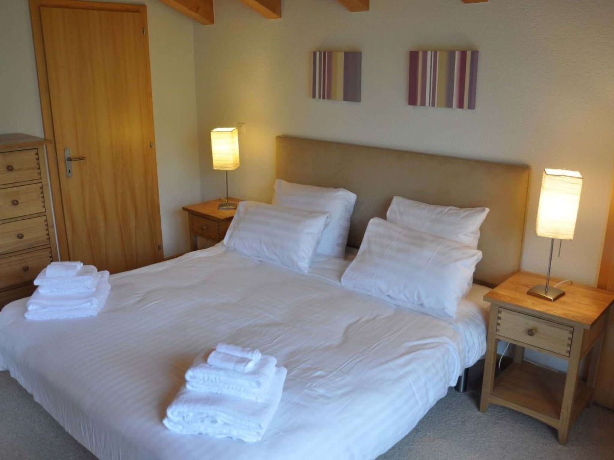 A neatly made bed with white linens and towels, two bedside tables with lamps, a wooden wardrobe, and a door.