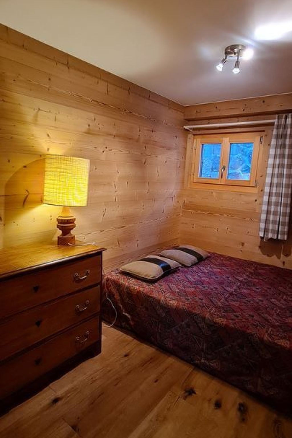 A room with wooden walls, a bed with patterned bedding, a bedside lamp, a window with curtains, and a wooden dresser.
