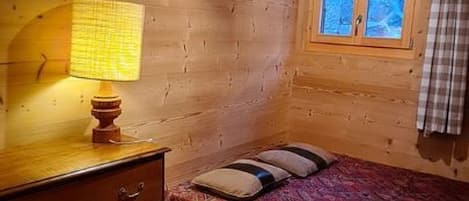 A room with wooden walls, a bed with patterned bedding, a bedside lamp, a window with curtains, and a wooden dresser.