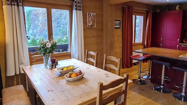 Un interior de madera con una mesa de comedor, sillas, un frutero, un florero con flores y una zona de cocina con barra.