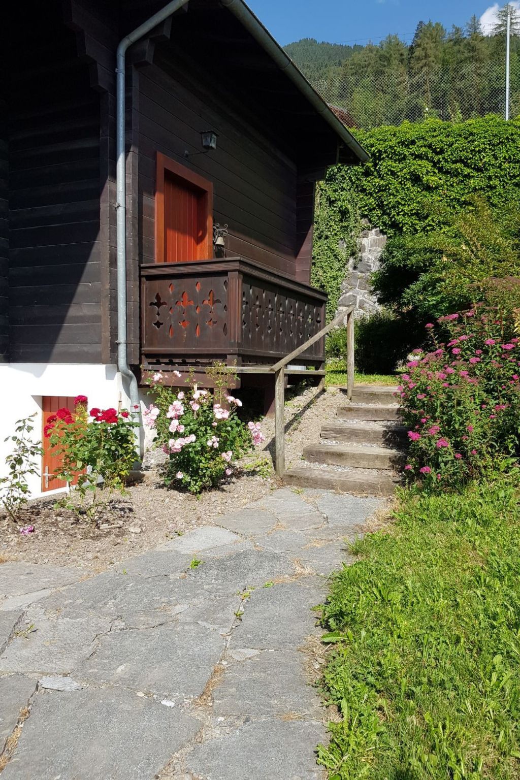 A wooden cabin with a stone staircase leading to a porch, surrounded by greenery and flowers.