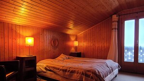 Habitación con paneles de madera, una cama, dos mesitas de noche, una silla y una ventana con cortinas.