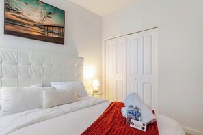 Ein Hotelzimmer mit Bett, weißem Kopfteil, einem Gemälde von einem Sonnenuntergang über einem Steg und einer weißen Tür.
