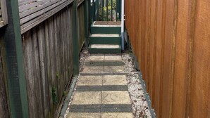 A narrow wooden pathway leading to a building with a 'Welcome' sign, surrounded by greenery and a clear sky.