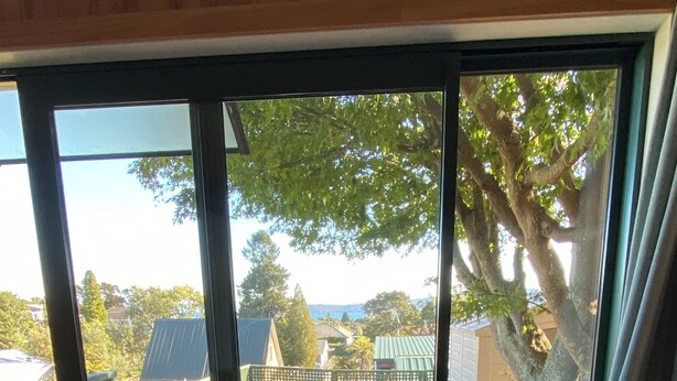 A room with a large window, a wicker sofa with a leaf-patterned pillow, and a wooden deck outside.