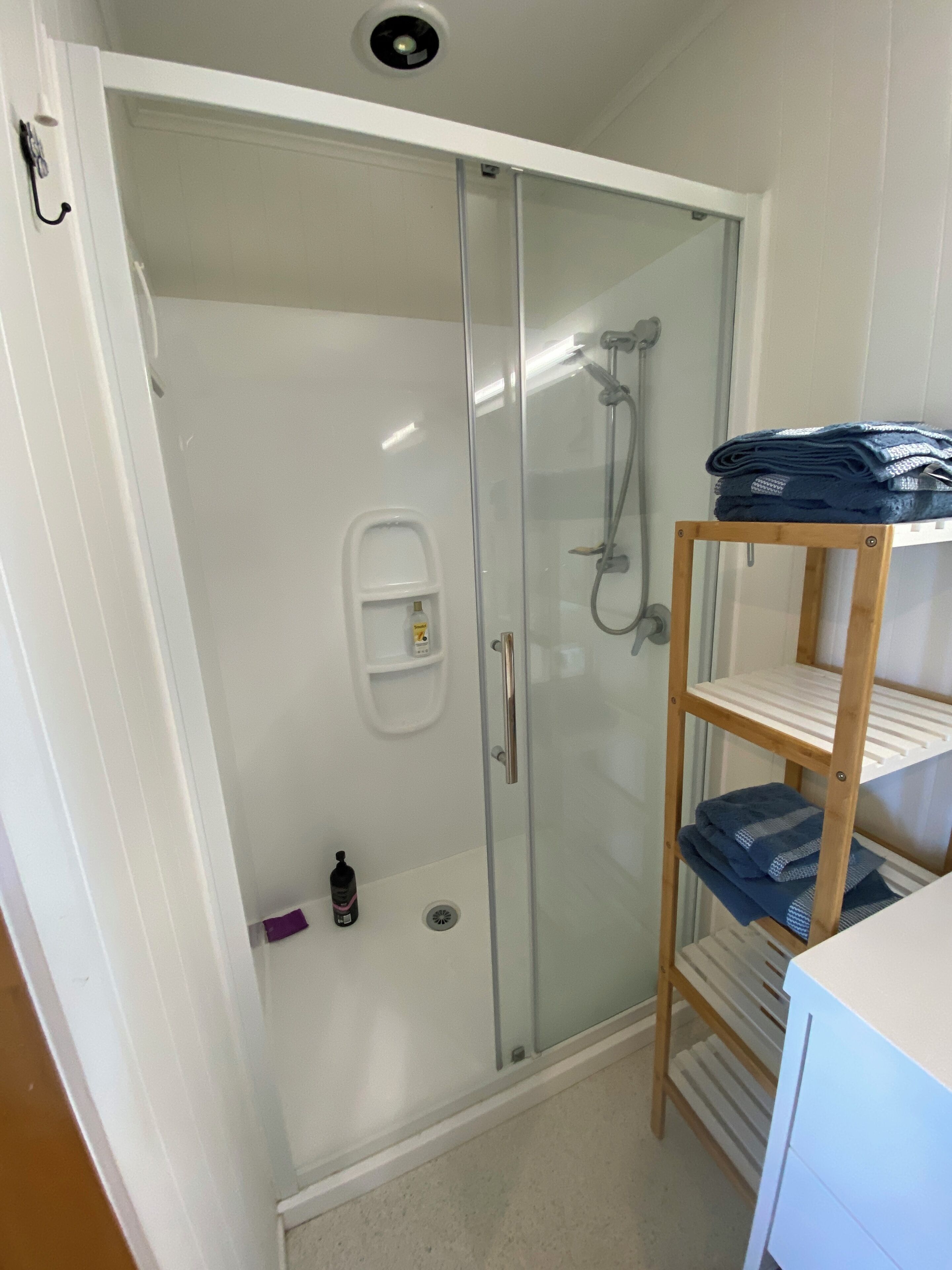 A walk-in shower with a glass door, a wooden shelf with folded towels, and a small shelf with toiletries.