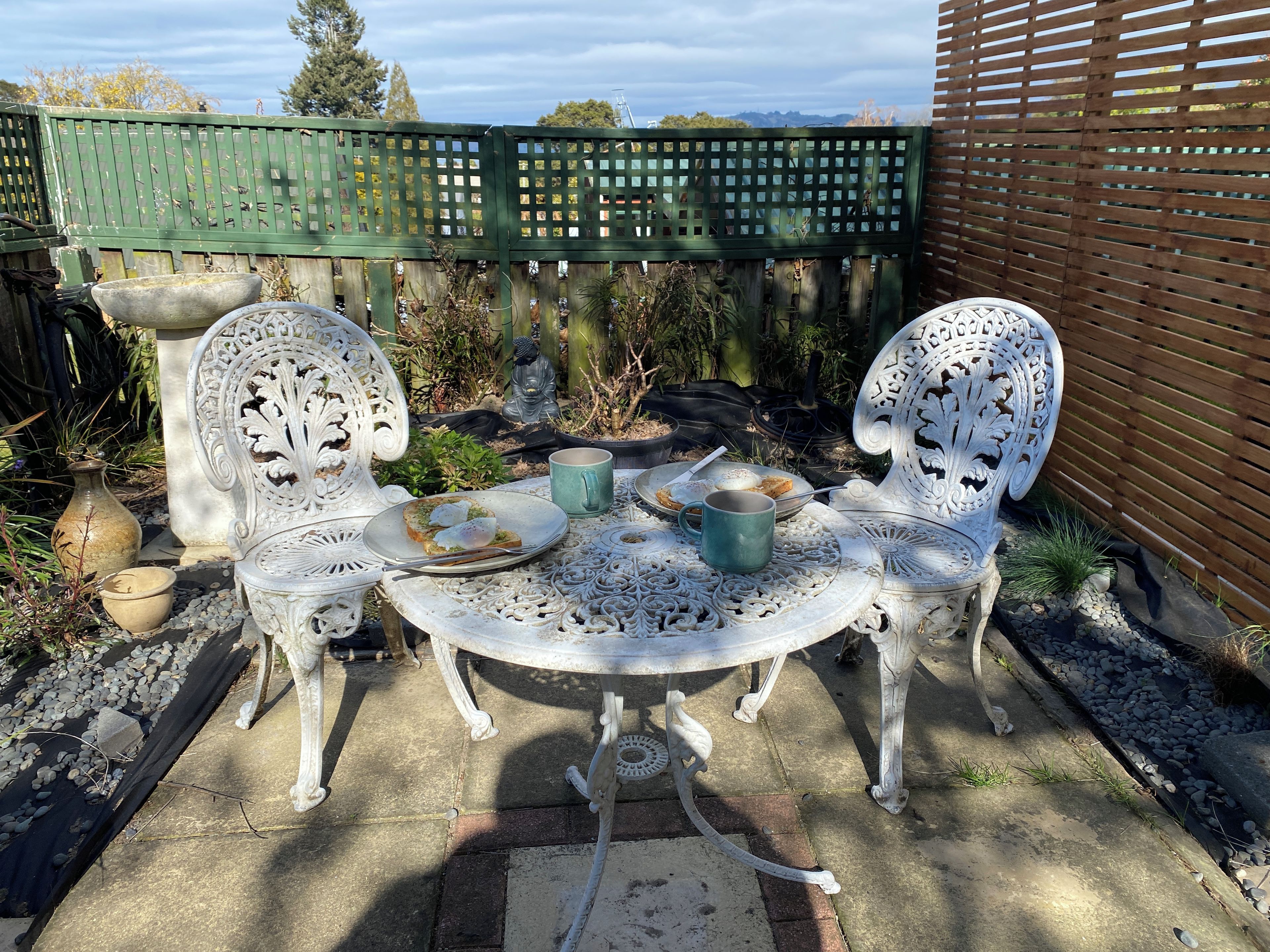 Un patio al aire libre con dos sillas blancas ornamentadas, una mesa redonda y una cerca verde al fondo.