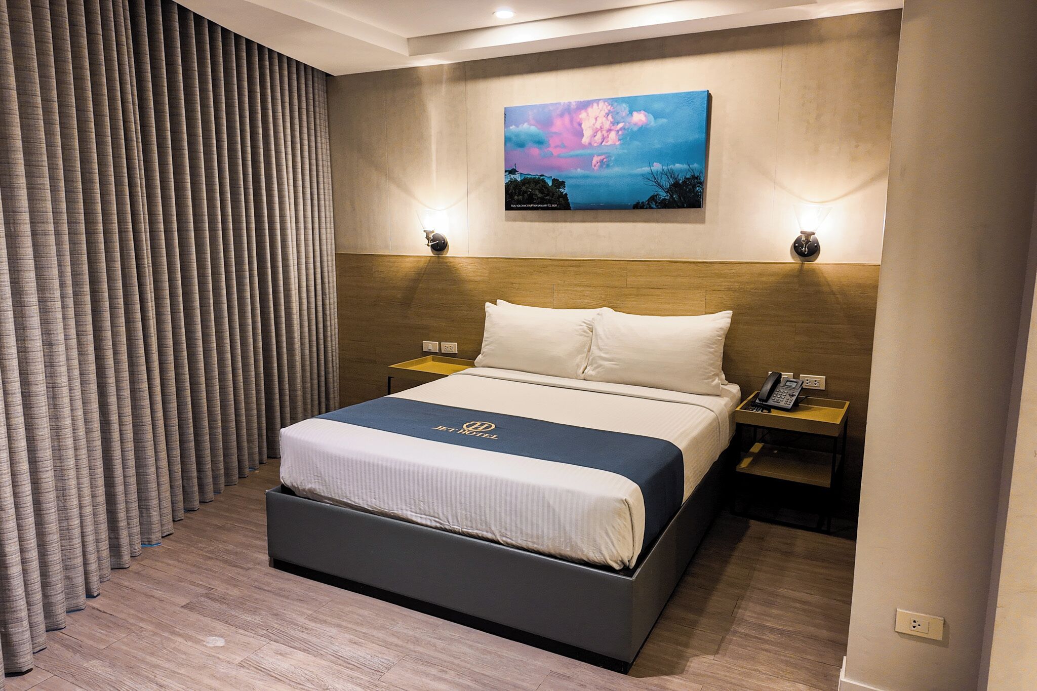 A hotel room with a large bed, a nightstand with a phone, a wall-mounted painting, and a curtain.