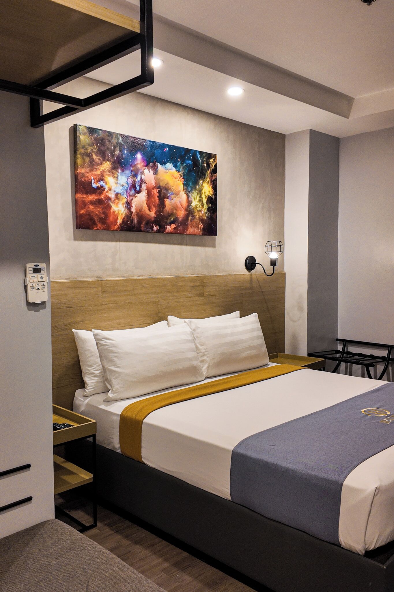 A modern hotel room with a large bed, a headboard, a wall-mounted artwork, a bedside table, and a nightstand.