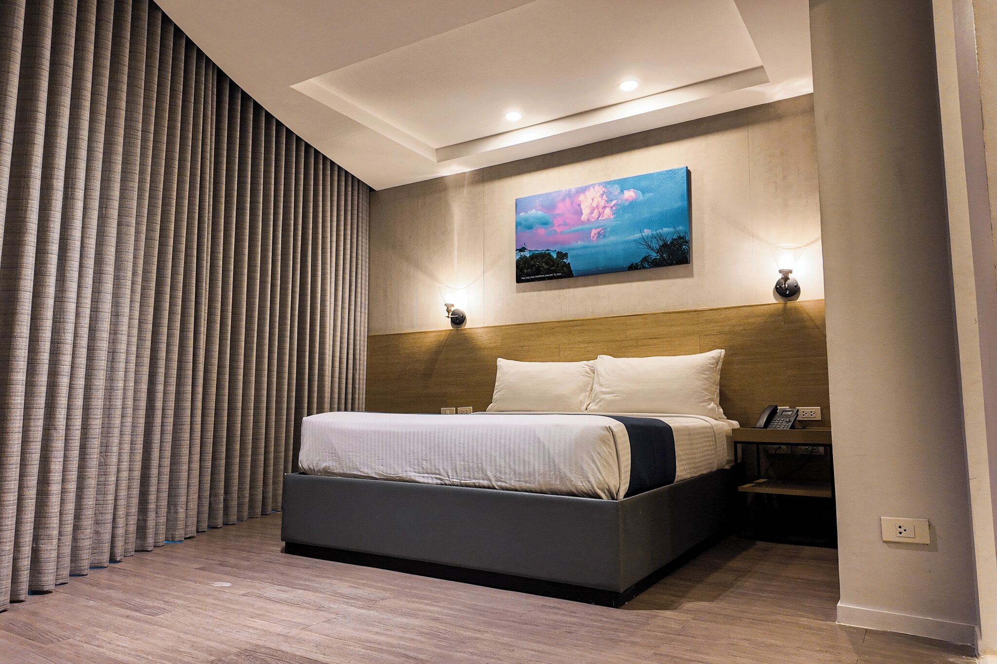 A modern hotel room with a large bed, a headboard with integrated lighting, a painting on the wall, and a nightstand with a phone.