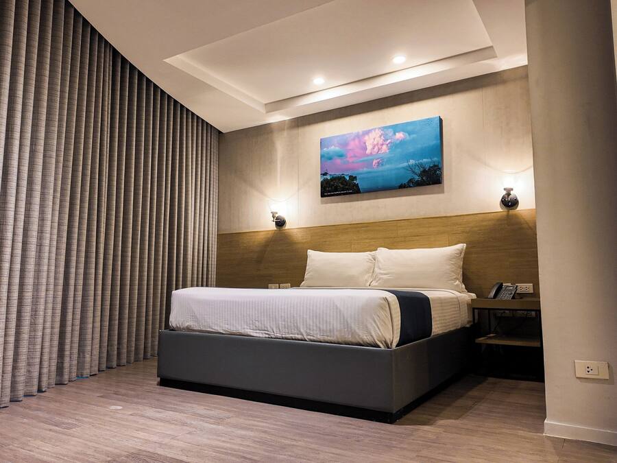A modern hotel room with a large bed, a headboard with integrated lighting, a painting on the wall, and a nightstand with a phone.