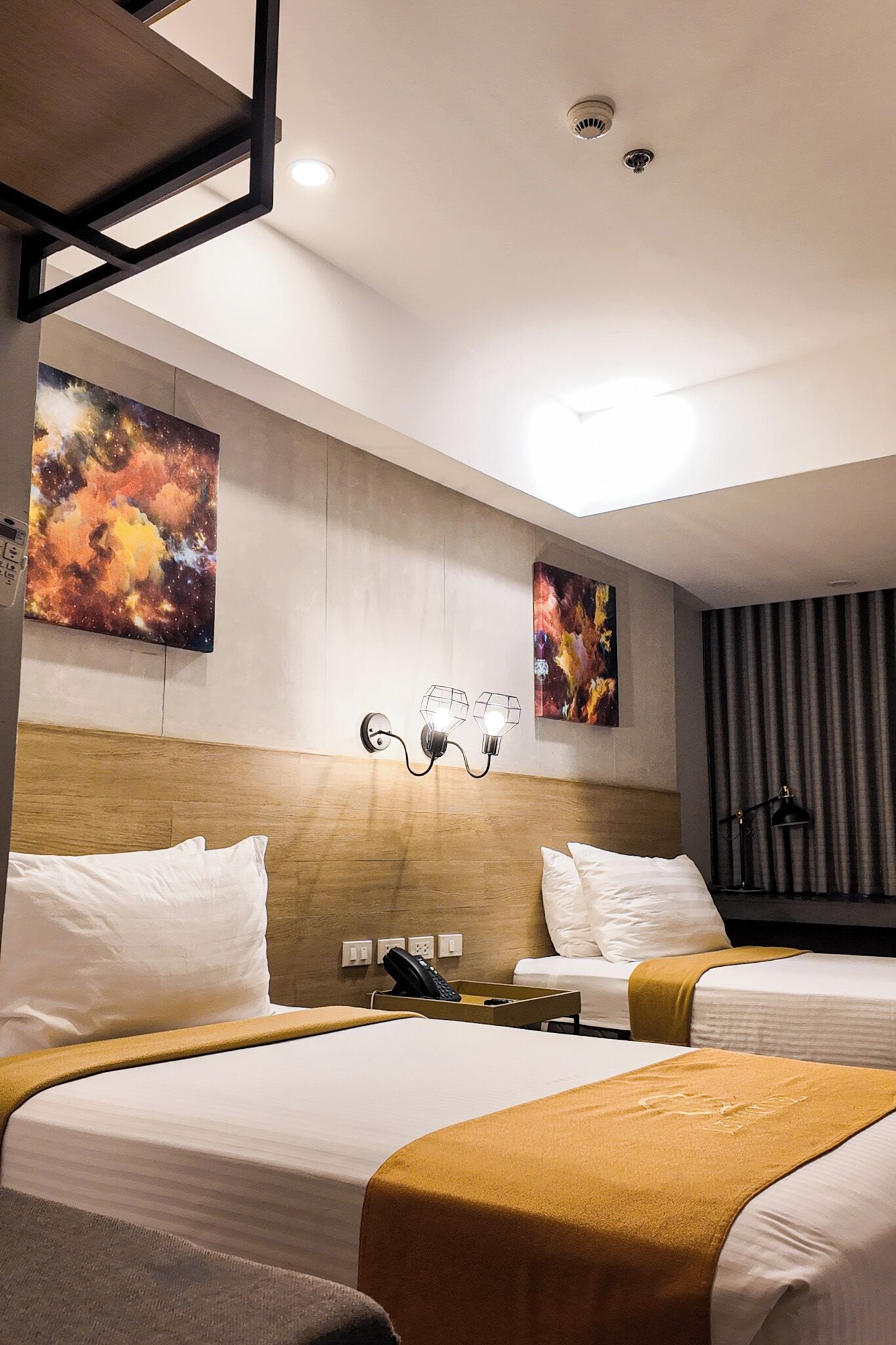 A hotel room with two beds, a headboard, artwork on the wall, a desk with a lamp, and a telephone.