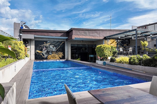 Una piscina con un mural, rodeada de follaje y un edificio moderno.