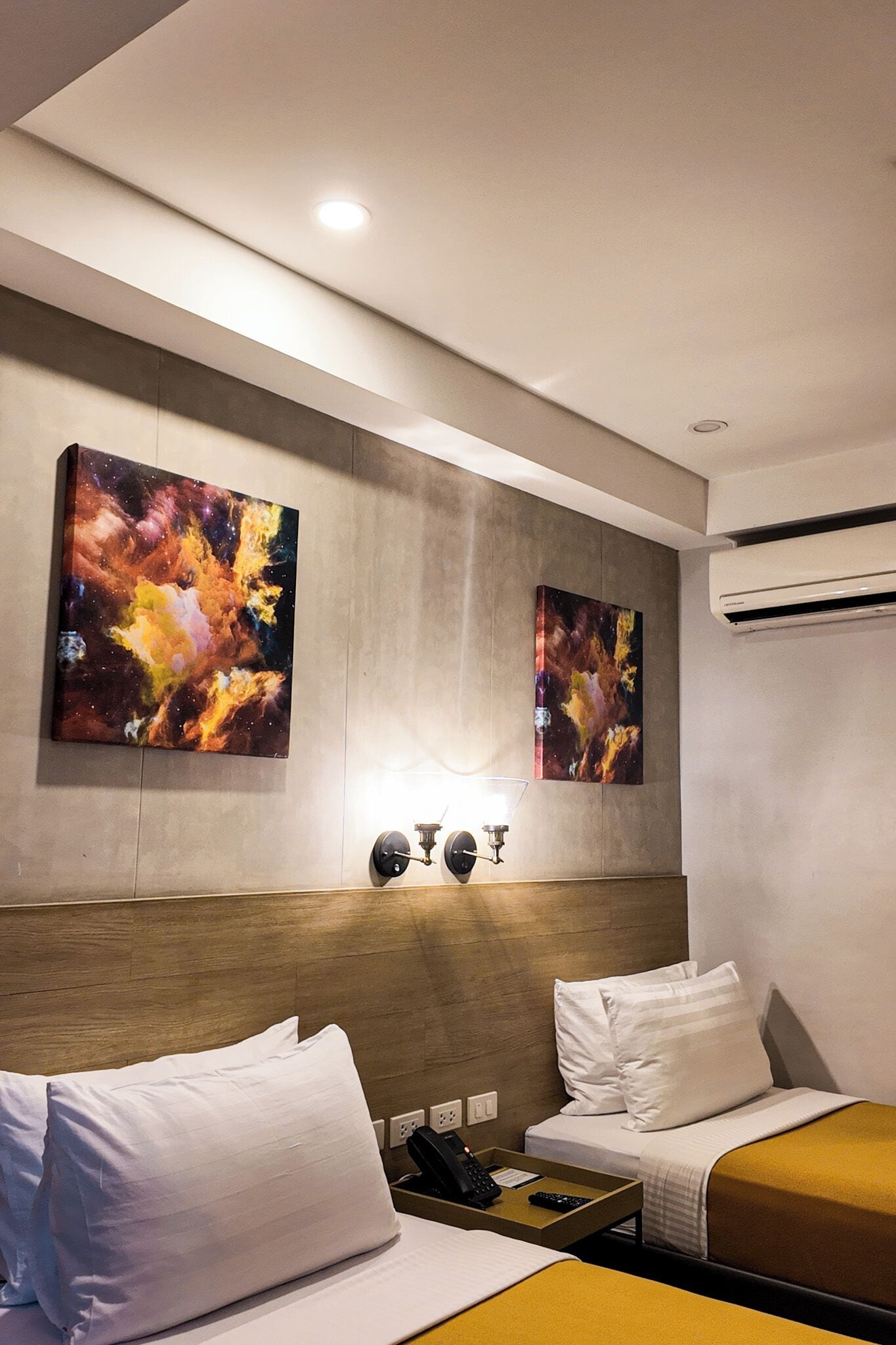 Habitación de hotel con dos camas, cabecera de madera, aire acondicionado de pared y dos cuadros abstractos.