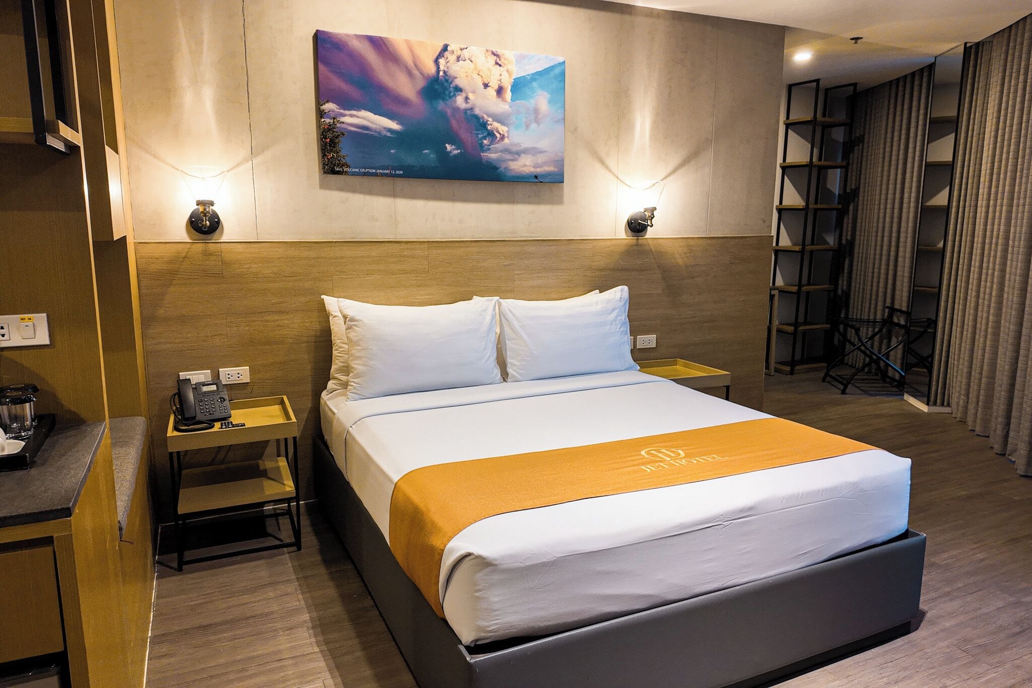 Una habitación de hotel con cama, mesitas de noche, teléfono, lámpara, un cuadro y cortina.