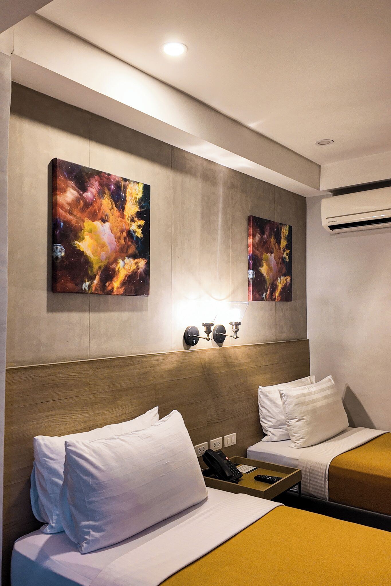 A hotel room with two beds, a wooden headboard, artwork on the wall, a bedside table with a phone and remote, and an air conditioning unit.