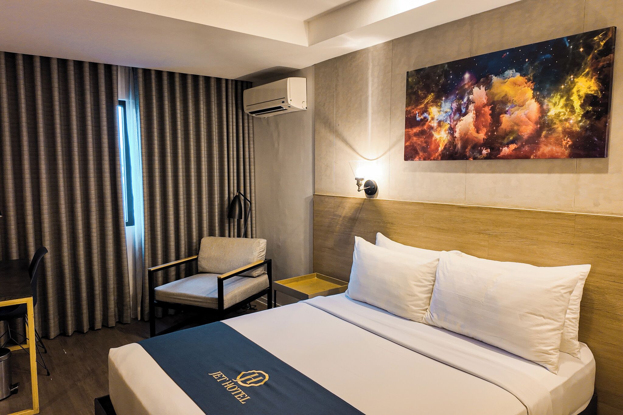 Habitación de hotel con una cama, una silla, un escritorio, aire acondicionado, un cuadro y una mesita de noche.