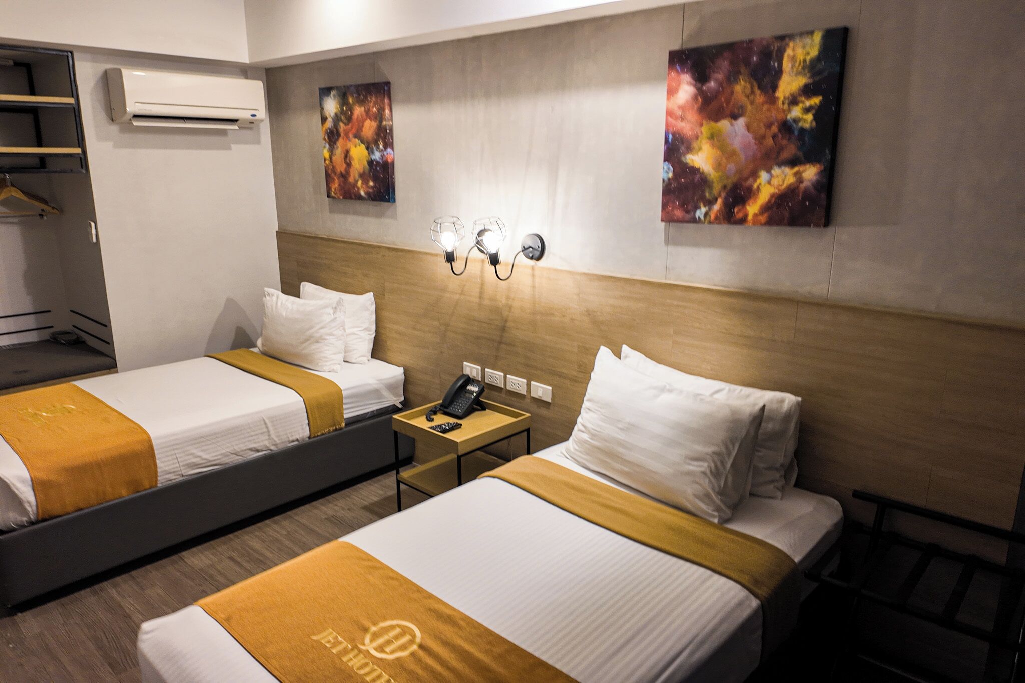 A hotel room with two beds, a bedside table with a phone, a wall-mounted air conditioner, and abstract paintings on the wall.