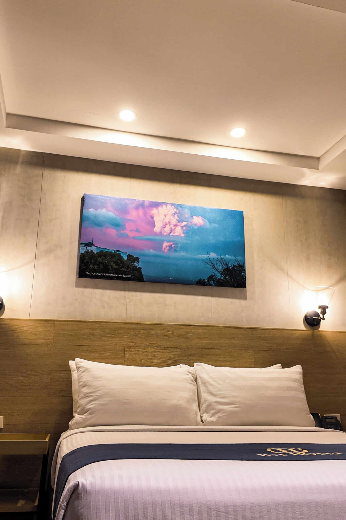A hotel room with a bed, a nightstand, a wall-mounted painting, and a ceiling light fixture.