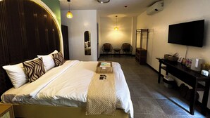 Quarto de hotel com cama, televisão, escrivaninha e área de estar com cadeiras.