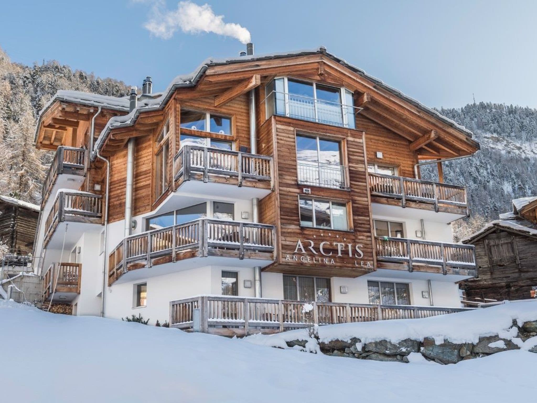 A multi-story wooden chalet with balconies, set against a snowy mountainous backdrop.