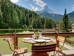 A wooden deck with a table set for two, overlooking a mountainous landscape with a clear sky. - Chalet Altesse, (Zermatt), 1054.01 by Interhome (Zermatt)