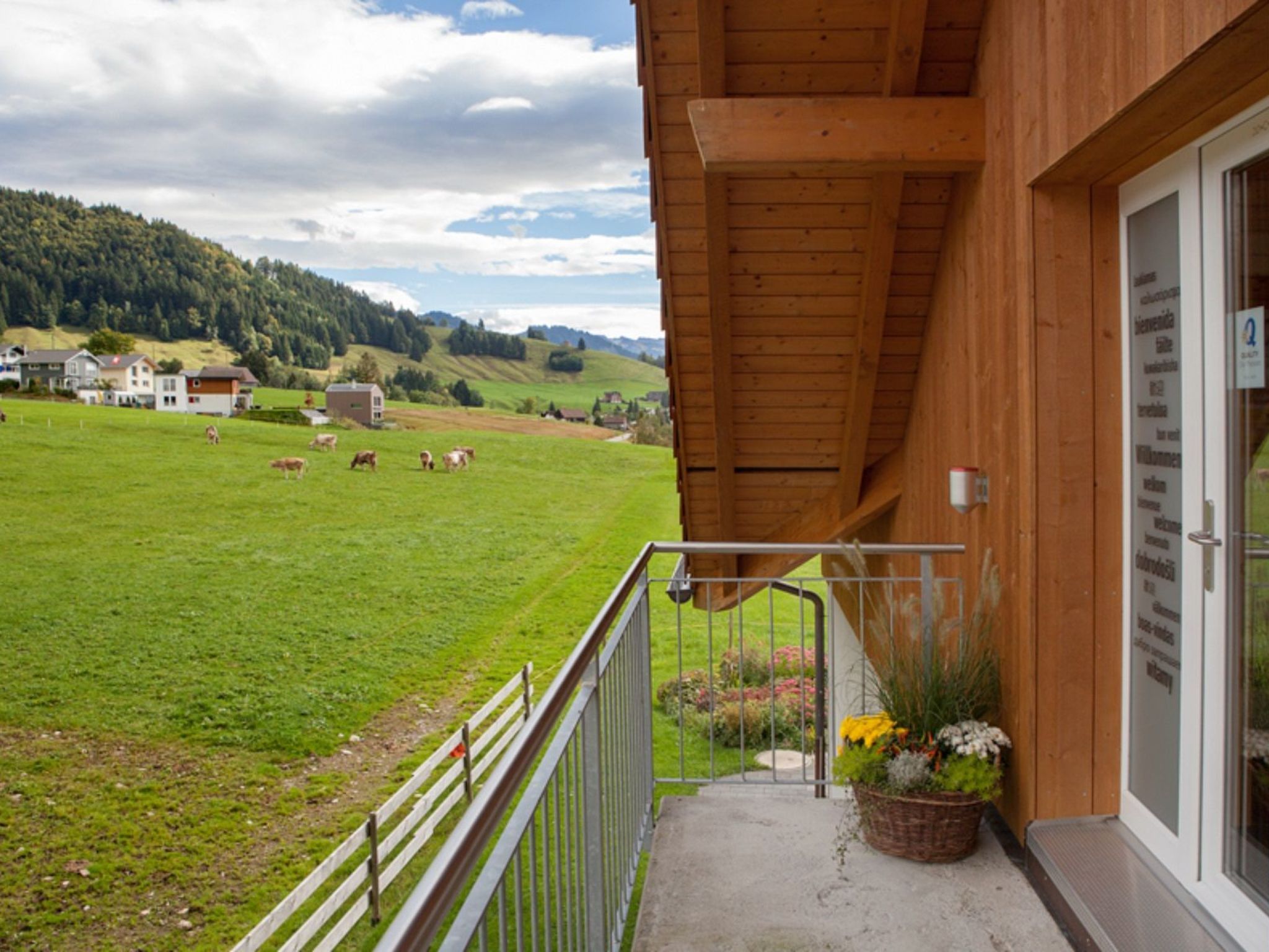 Varanda de madeira com vista para um campo verde e vacas, uma porta branca e uma placa com texto.