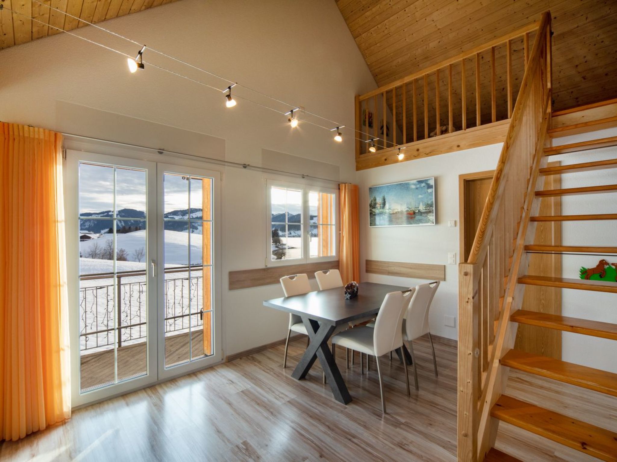 A spacious dining area with a table and chairs, large windows with a view of a snowy landscape, and a wooden staircase.