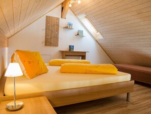 Ein Schlafzimmer im Loft-Stil mit schrägem Dach, einem Bett mit gelber Bettwäsche, einem Nachttisch, einer Lampe und einem kleinen Holzboden mit Büchern.