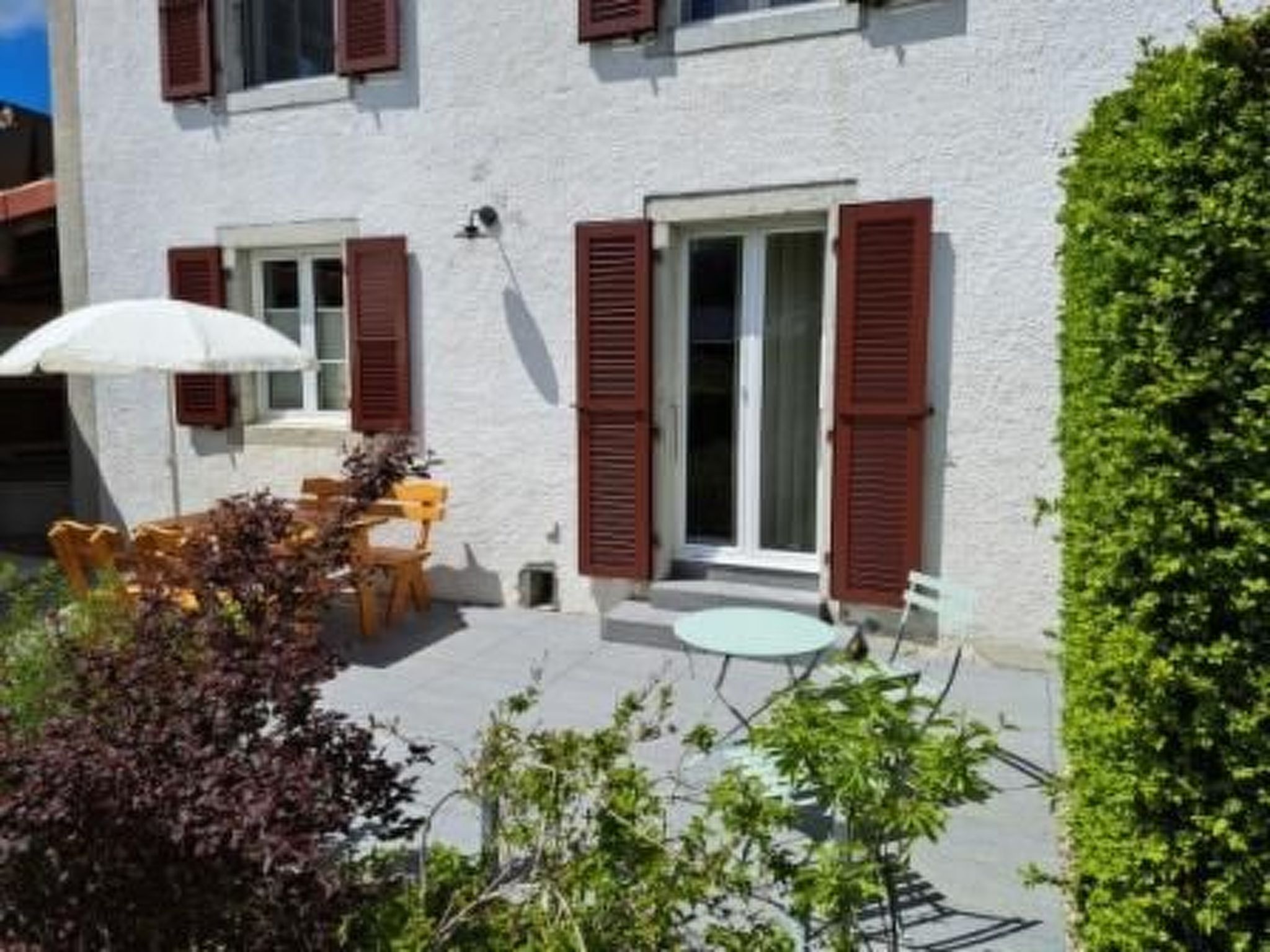 Un bâtiment à l’extérieur blanc, avec des volets rouges, un petit patio doté d’une table et de chaises, et un parasol blanc.