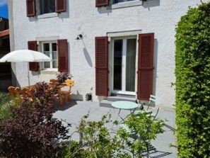 A building with a white exterior, red shutters, a small patio with a table and chairs, and a white umbrella. - Appartement - Le Pèlerin, (Saignelegier), by Interhome (Saignelegier)