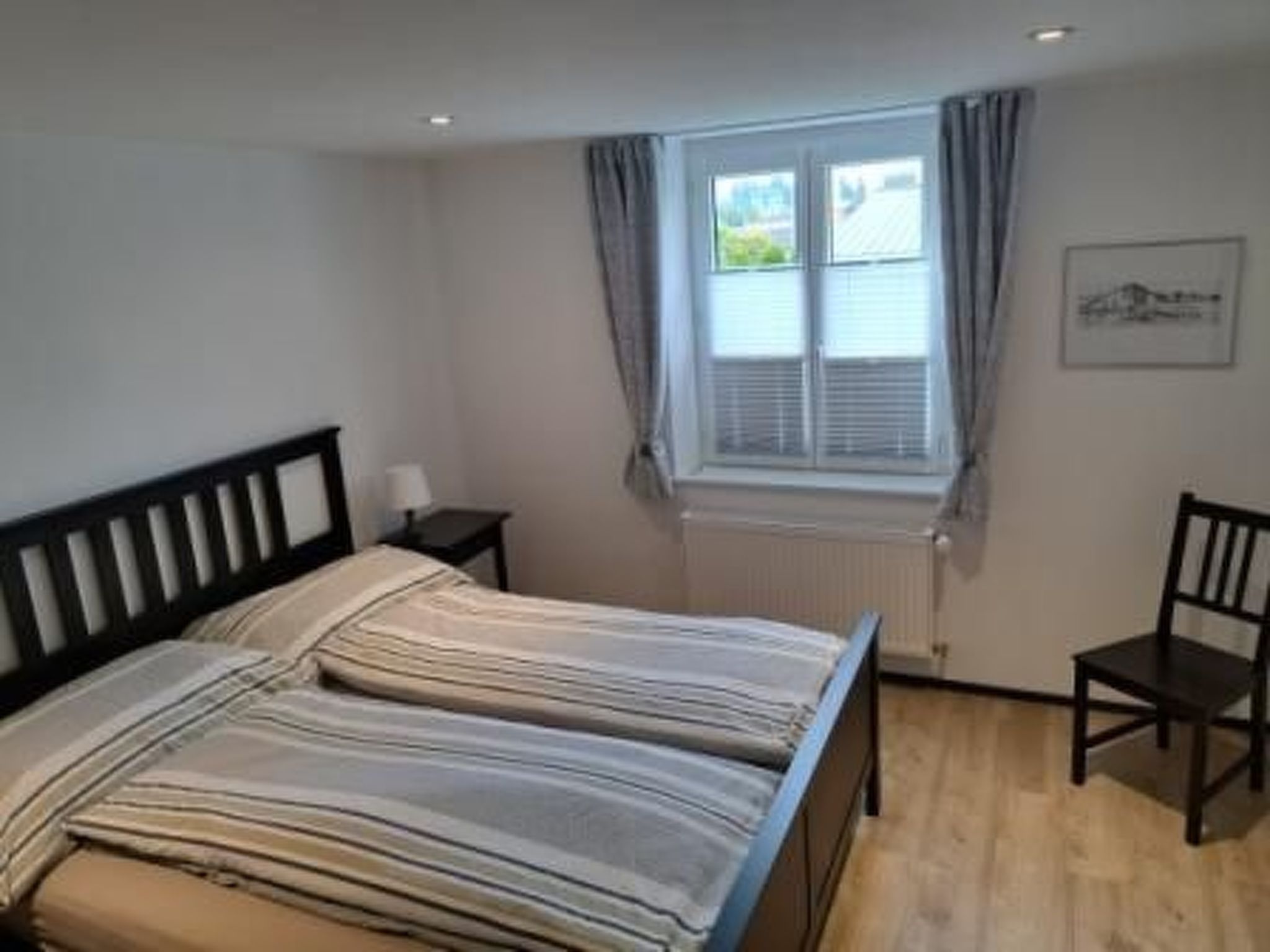 Ein Schlafzimmer mit einem Doppelbett, einem Sessel, einem Nachttisch, einem Fenster mit Vorhängen und einer Heizung.