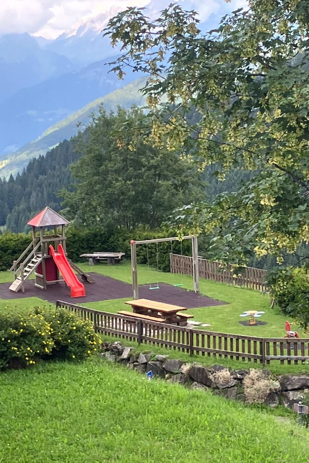 Une aire de jeux avec un toboggan et des balançoires, entourée d’une clôture en bois et d’un mur de pierres.