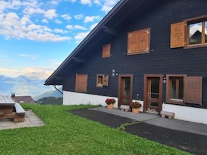 A two-story building with wooden shutters, a small porch with potted plants, and a view of mountains in the background. - Oeillet 4, (Champoussin), by Interhome (Les Crosets)