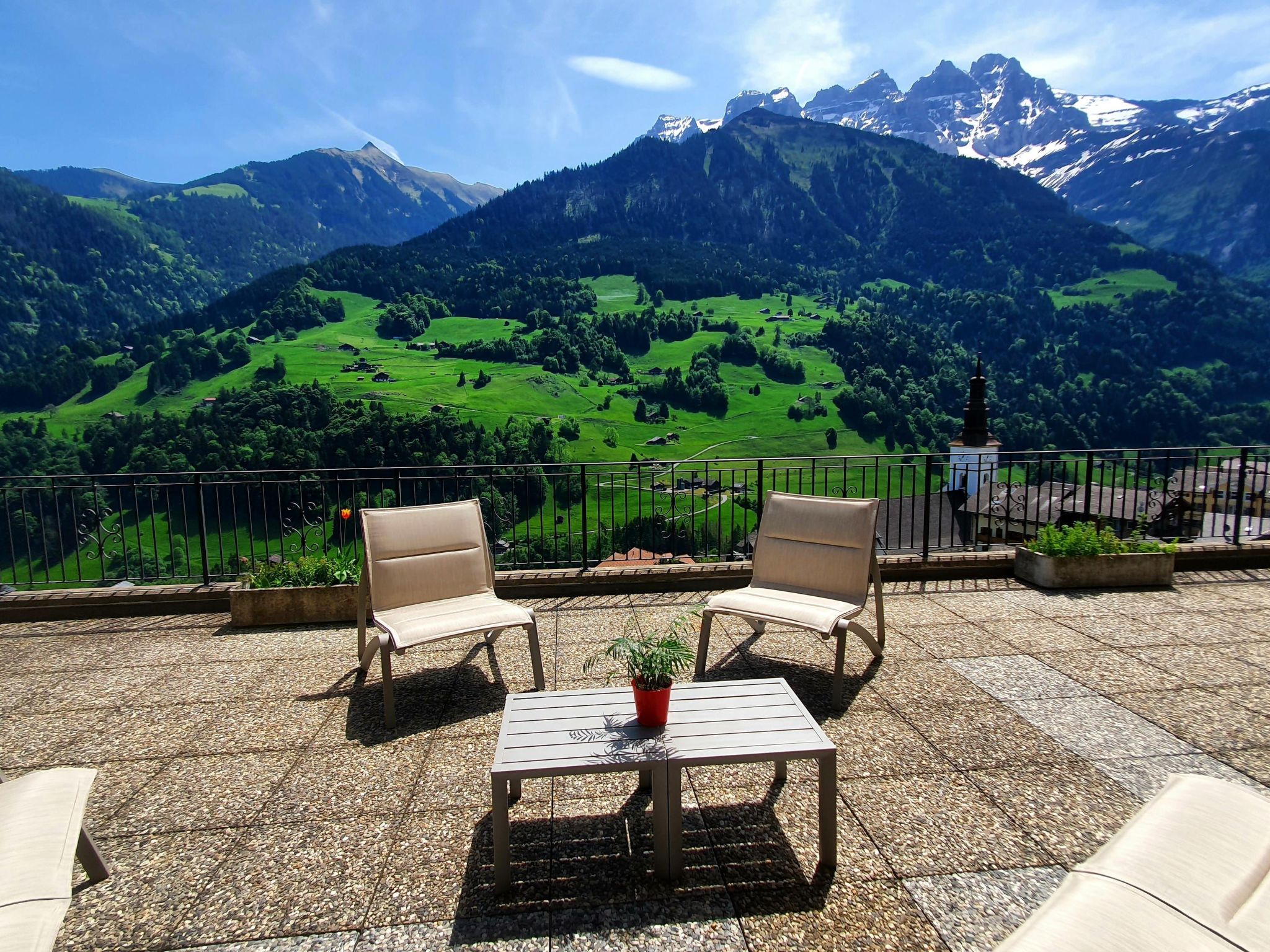 Une terrasse avec deux chaises et une table, d’où l’on peut admirer un paysage montagneux avec des champs verdoyants et le clocher d’une église.
