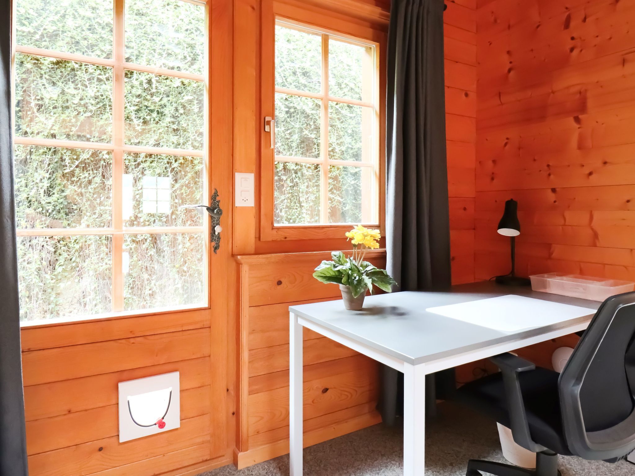 A wooden room with a white desk, a chair, a potted plant, and a window with curtains.