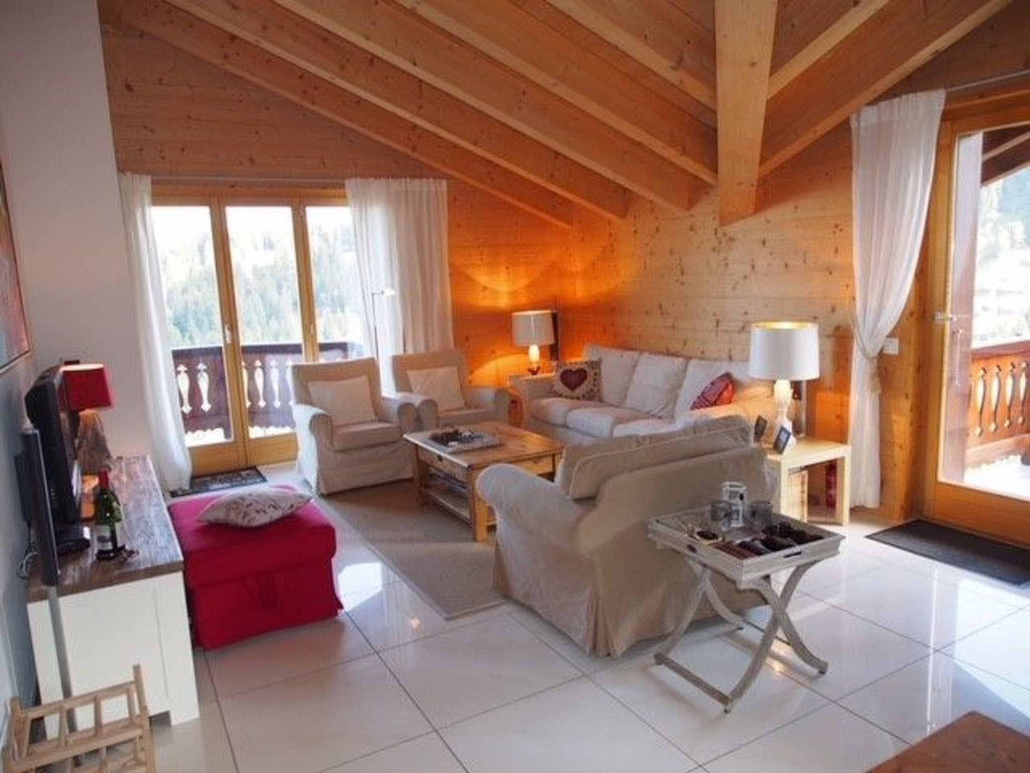 Uma sala de estar aconchegante com sofá, poltronas, mesa de centro e um banquinho vermelho. Há uma televisão à esquerda e uma janela com cortinas.