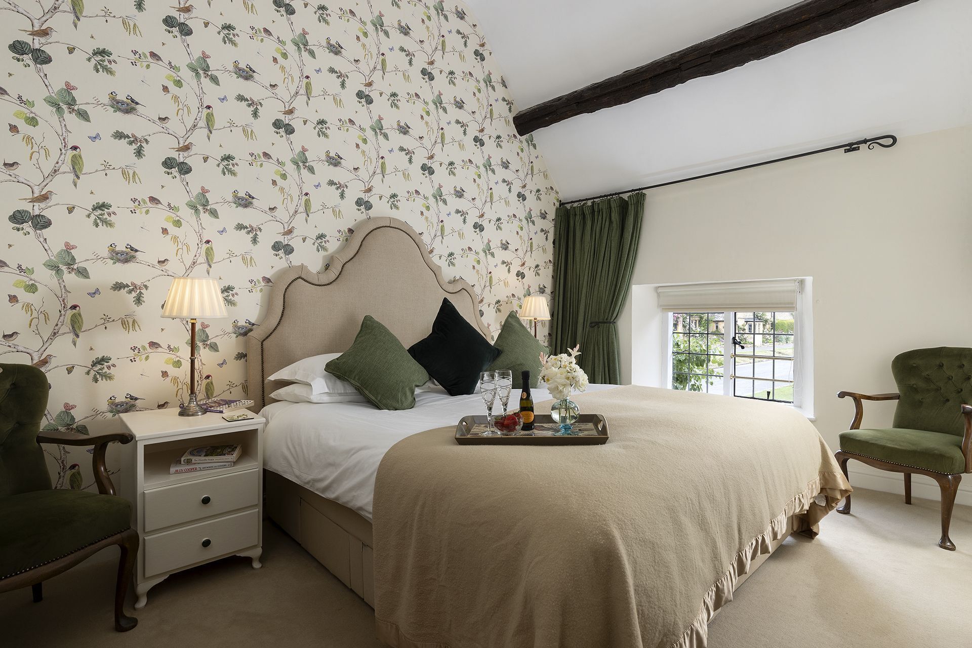 A bedroom with a floral wallpaper, a bed with a beige headboard, a nightstand with a lamp, a chair, and a window with curtains.