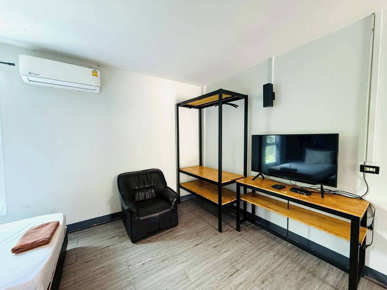 Una habitación con una cama, un sillón de cuero negro, un televisor sobre un soporte de madera y un estante metálico negro.