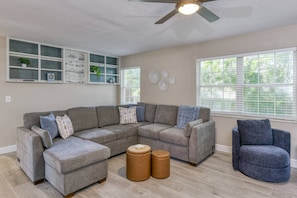 A living room with a sectional sofa, a blue armchair, two ottomans, a ceiling fan, and a window with blinds. - Short Walk to the Beach, Screened Porch, Renovated Home, 4 miles to Downtown, $300 in Free Tickets! (St. Augustine)
