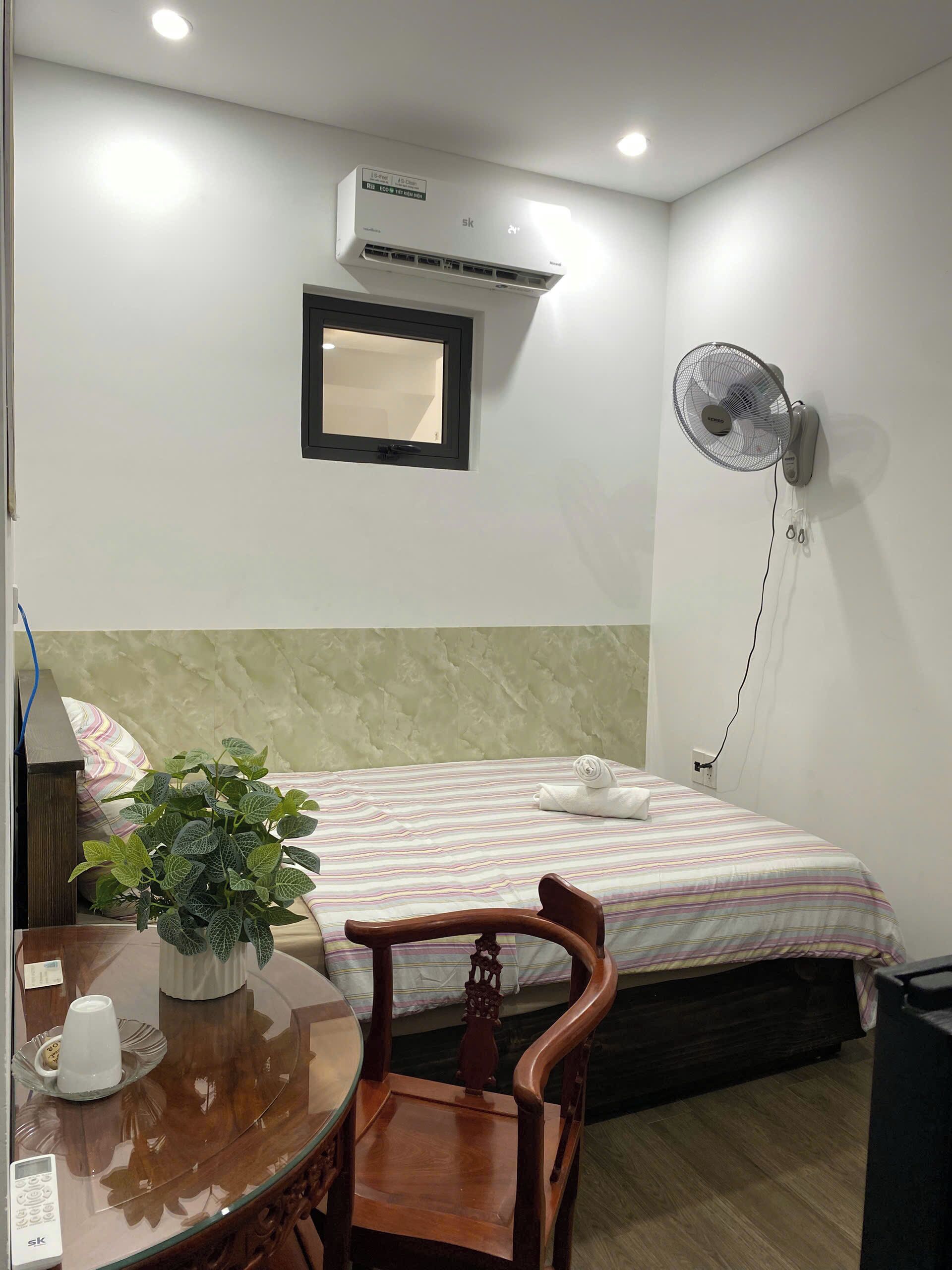 A hotel room with a bed, bedside table, chair, potted plant, and a wall-mounted air conditioner.