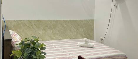 A hotel room with a bed, bedside table, chair, potted plant, and a wall-mounted air conditioner.
