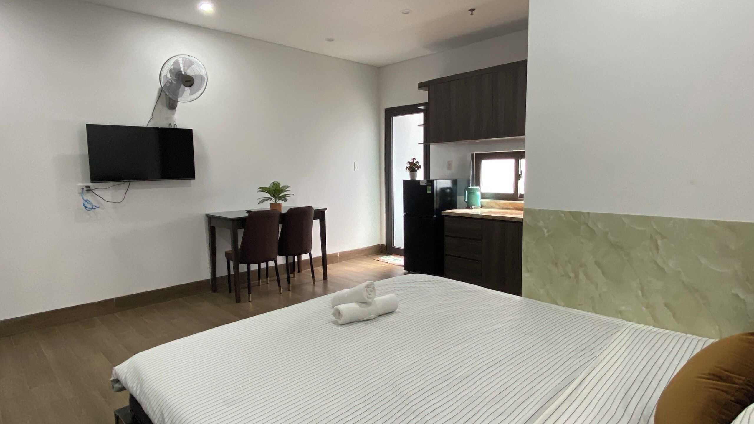 A hotel room with a bed, a desk with a chair, a television mounted on the wall, and a kitchen area visible through an open door.