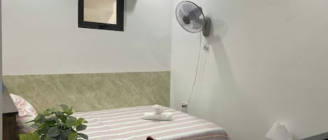 A hotel room with a bed, a desk, a chair, a potted plant, an air conditioner, a wall-mounted fan, and a lamp.