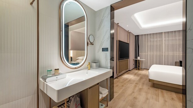 A modern hotel room with a large bed, a TV, a bathroom with a vanity, and a mirror.