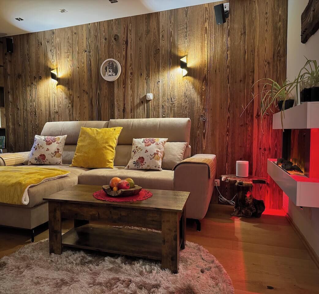 A cozy living room with a wooden wall, a sofa with floral cushions, a coffee table with fruit, and a bookshelf.