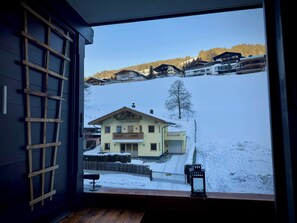 Blick durch ein Fenster auf ein Gebäude mit Holzgitterwerk, einem Balkon und einer verschneiten Landschaft mit Häusern.