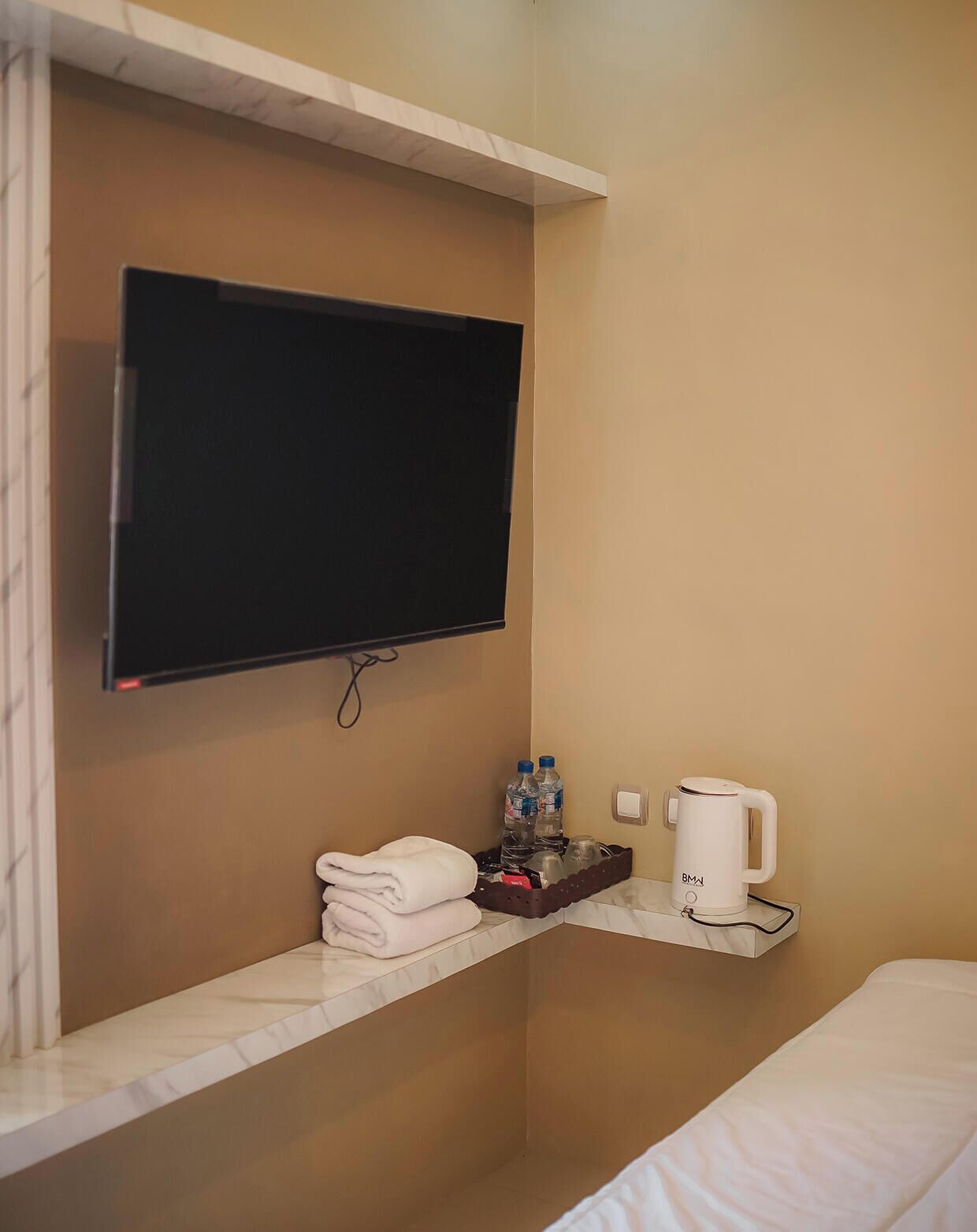 Una camera d'albergo con TV a schermo piatto fissata al muro, un piccolo ripiano con articoli da toilette e un letto.